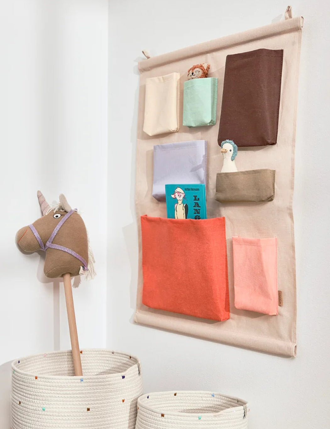 Wall-mounted fabric organizer with colorful pockets holding toys and books, next to woven baskets on a white wall.