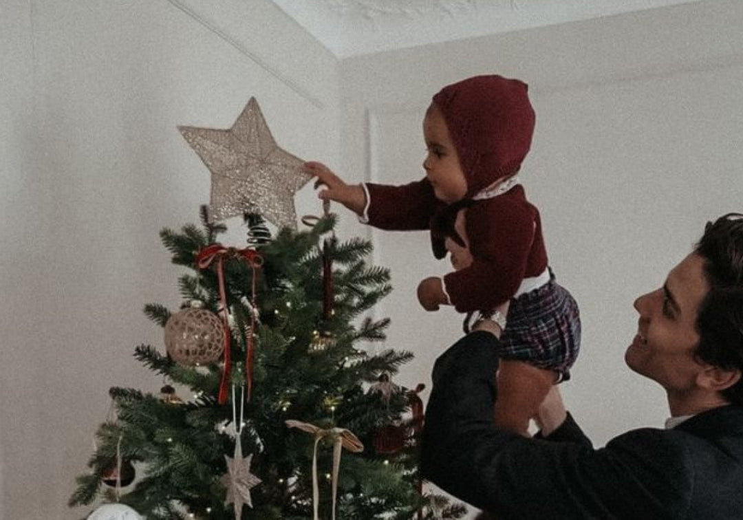A baby with a red bonnet, being lifted up by their dad to put a star on top of the christmas tree