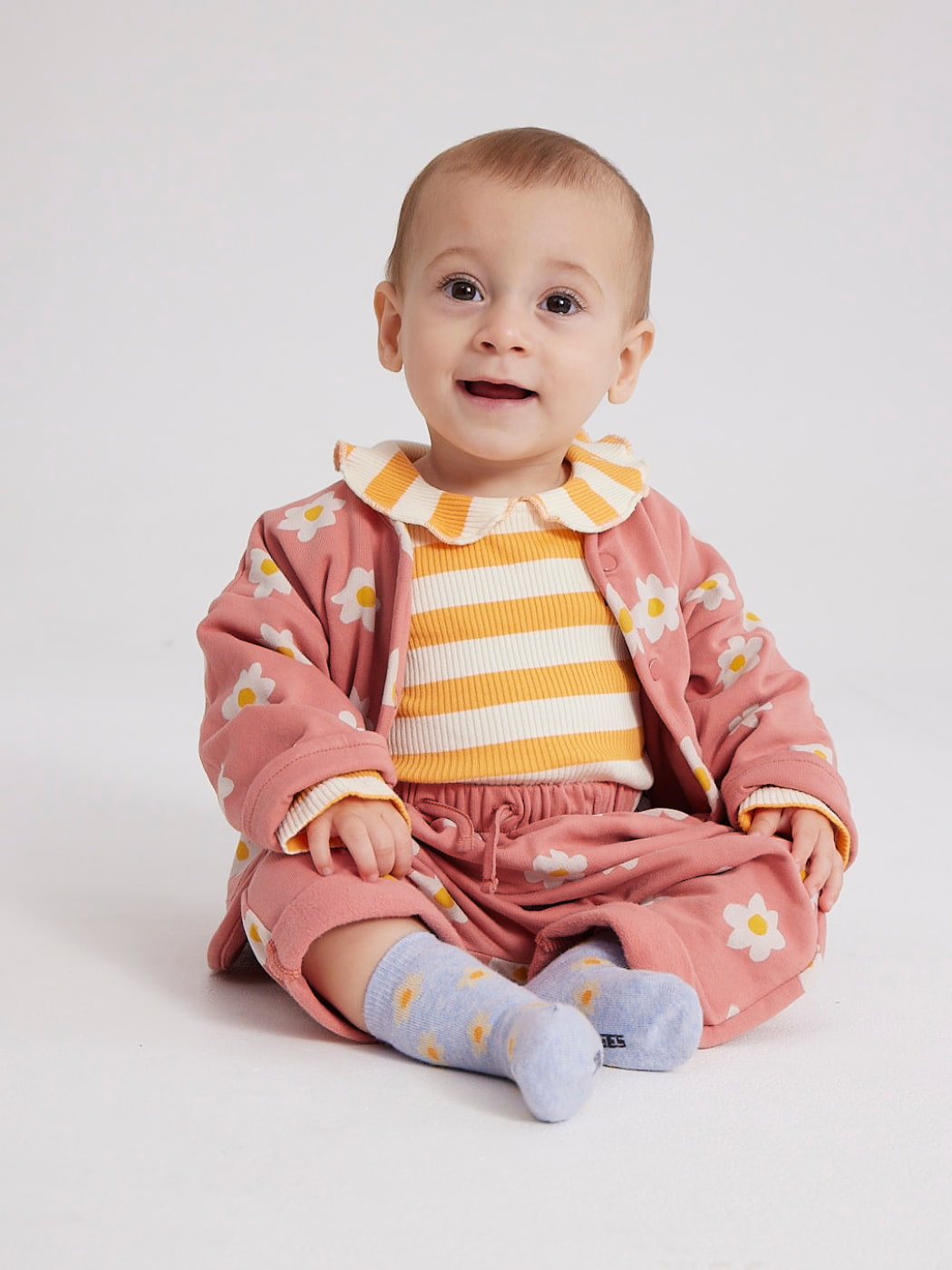 a baby sitting wearing an outfit by bobo choses including a button up pink jacket with flowers on it and a stripey yellow top underneath