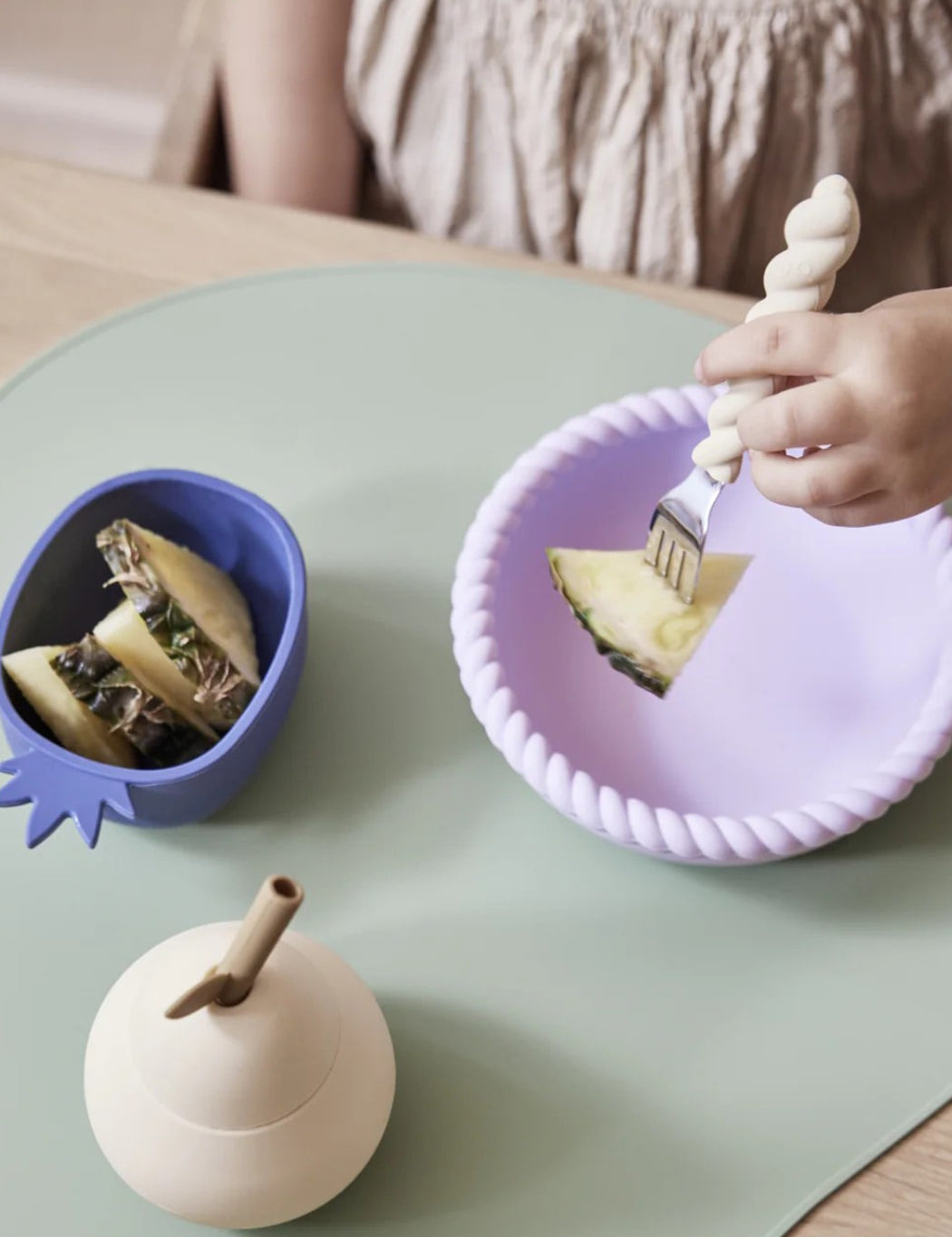 Child eating a slice of pineapple with a fork on a purple plate, next to a blue bowl with a sandwich on a light green table.